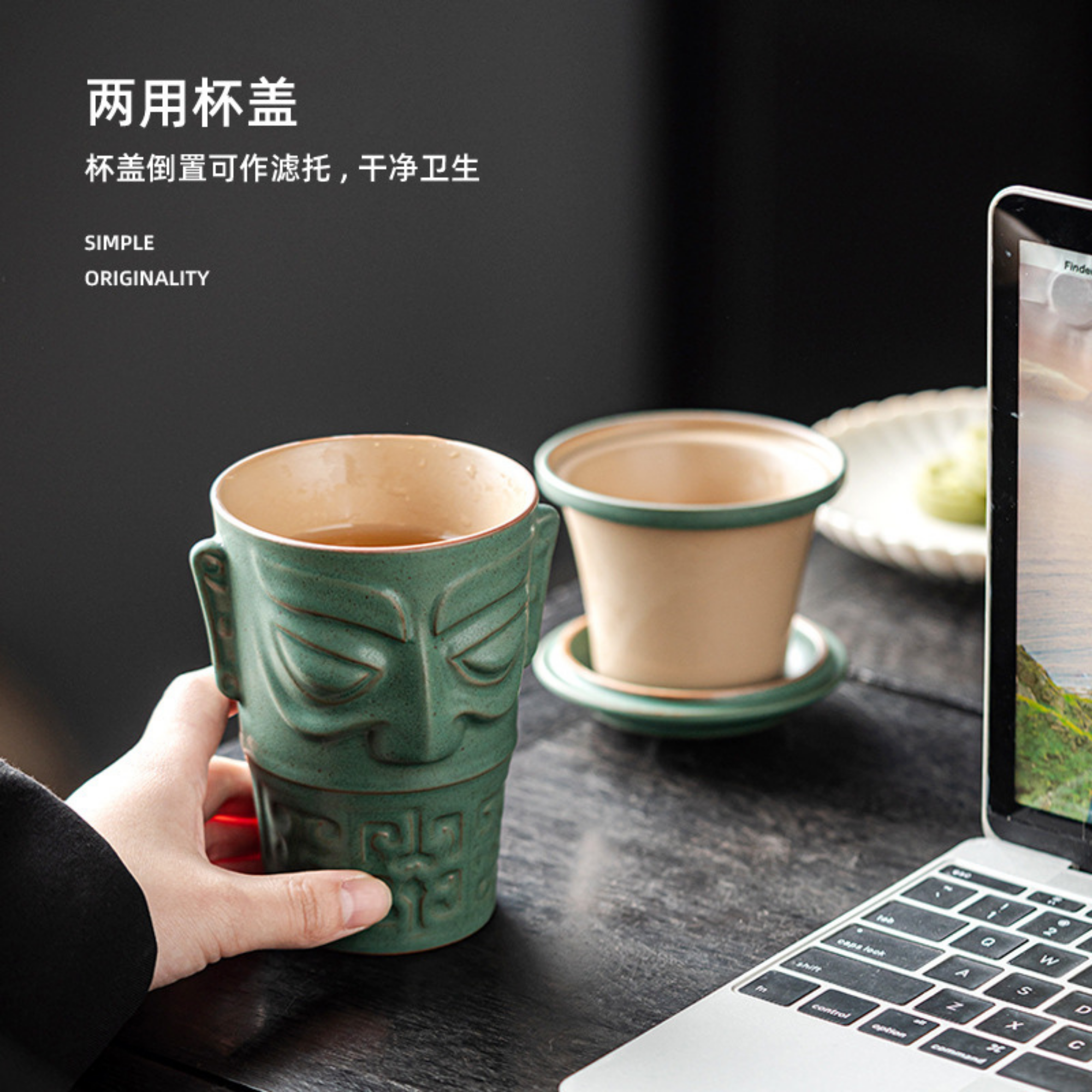 Green ceramic mug with face design held by a hand on a desk with a laptop.