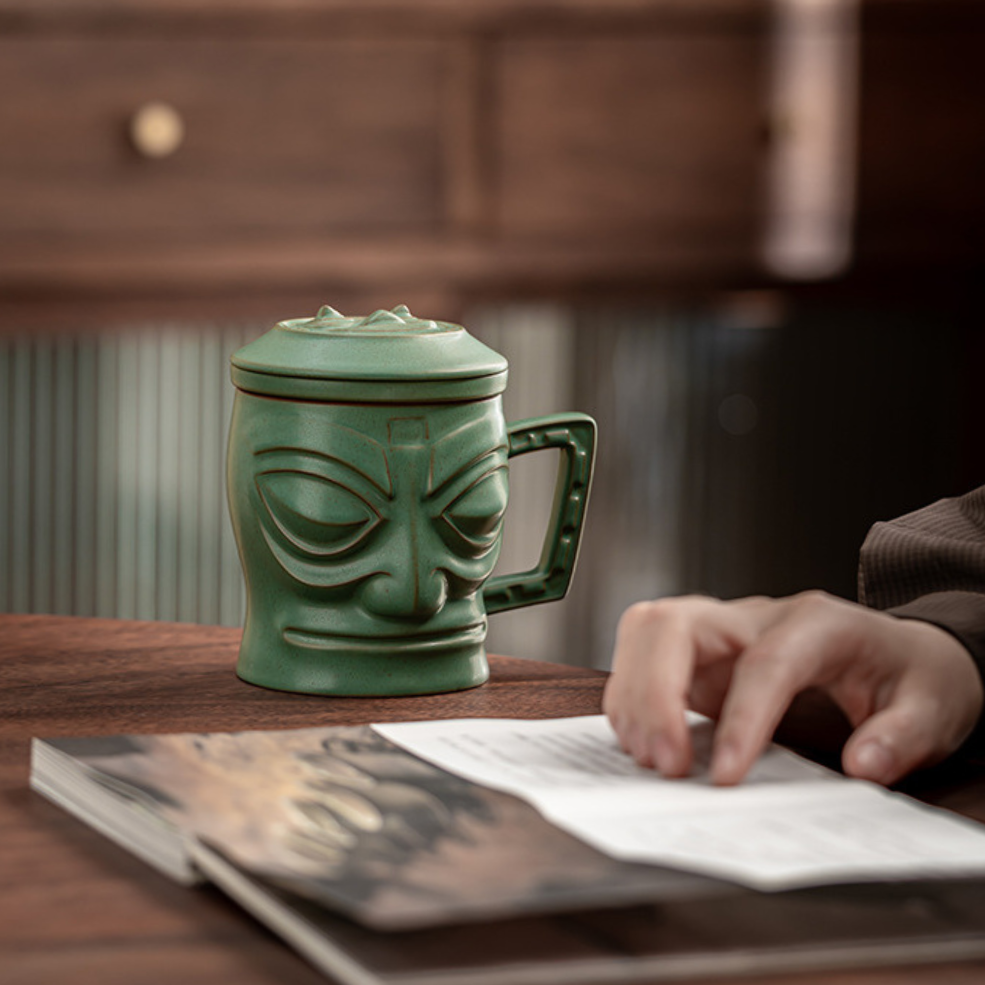Green tiki mug with a face design on a wooden table, with a person reading a book in the background.