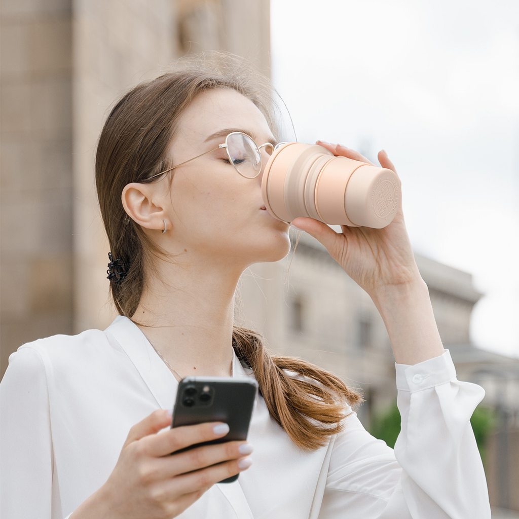 Woman drinking from a pink cup and holding a phone outdoors.
