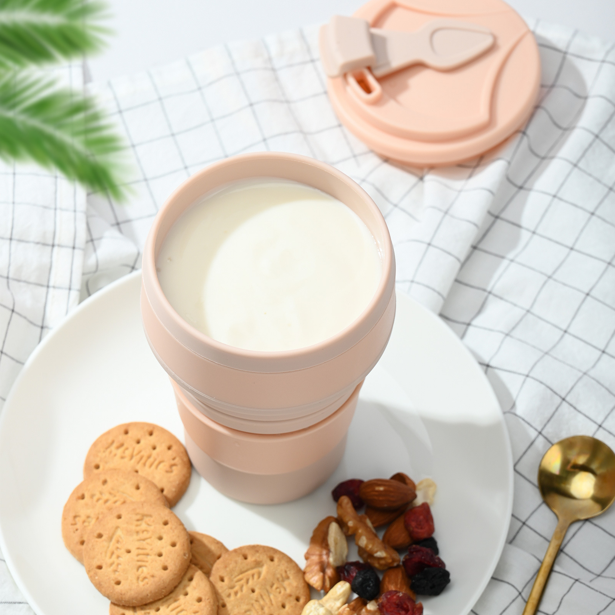 Pink collapsible cup with milk, cookies, and nuts on a white plate with a checkered tablecloth.